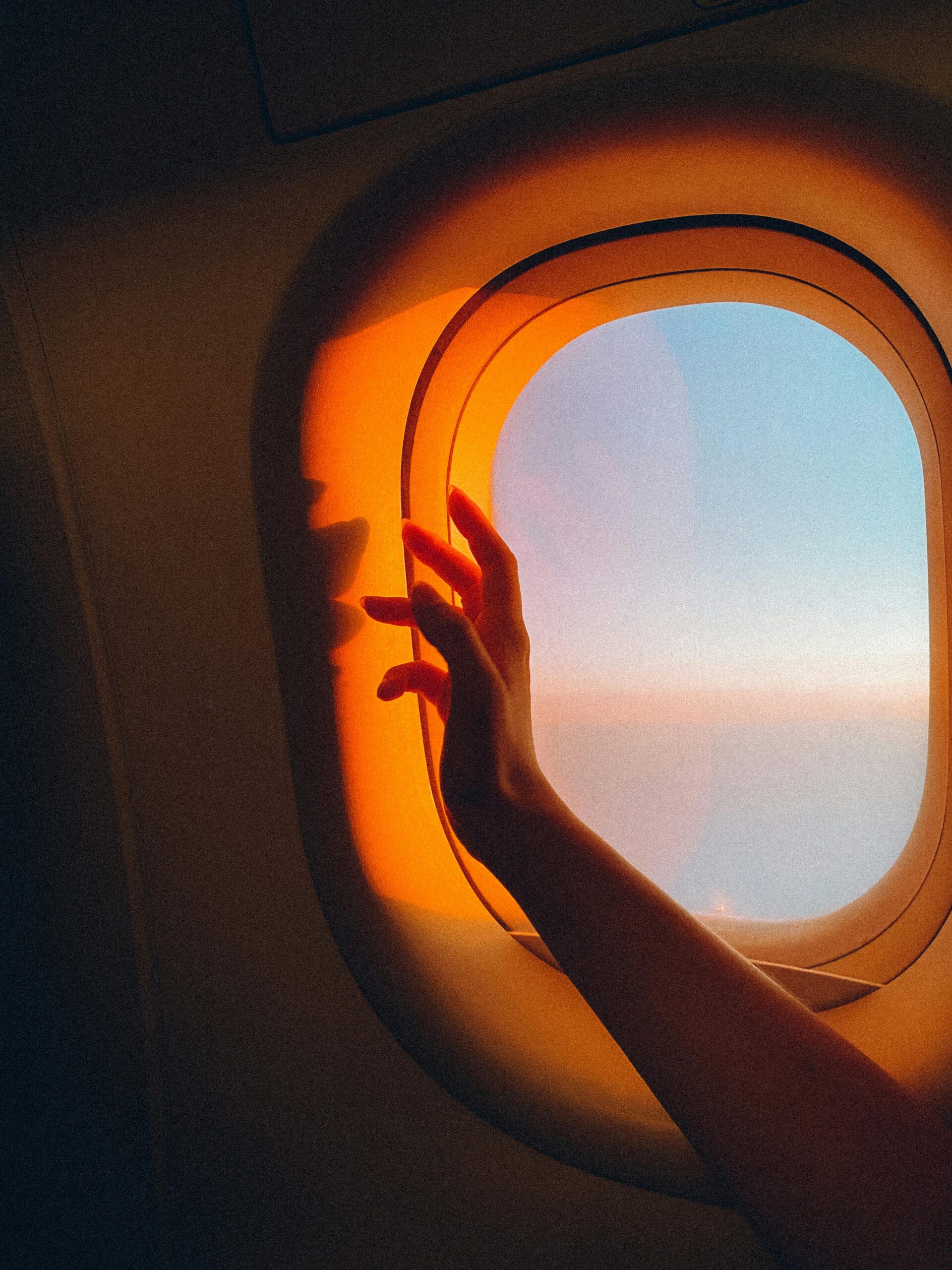 Hand reaches towards a sunset view from an airplane window, capturing a travel moment.